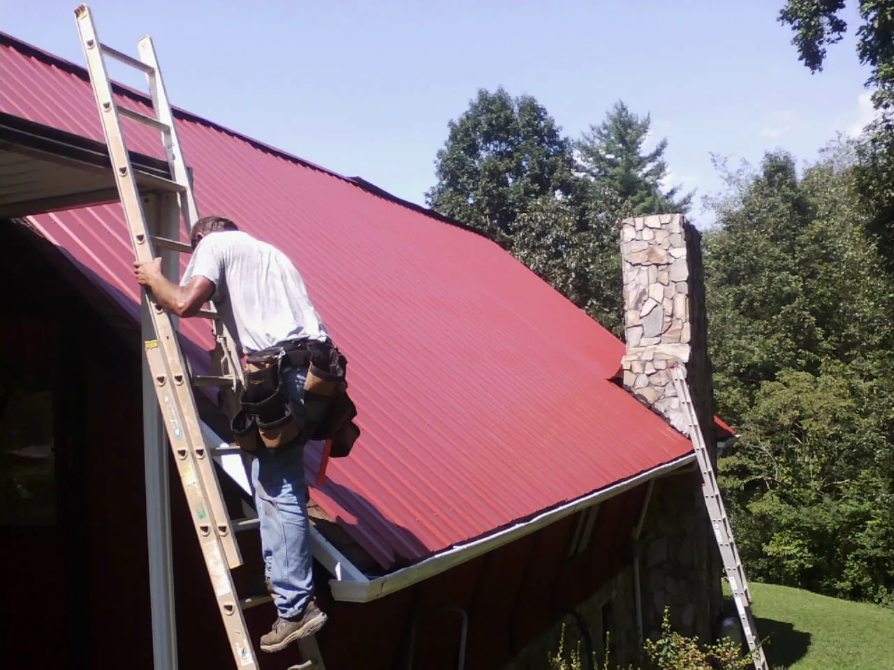 Roofer on a ladder beside a red metal roof and stone chimney — Soffit & Fascia Repair by Roofing Brothers in Browns Valley